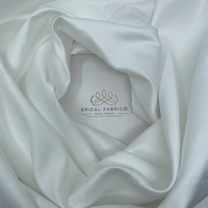 May include: White satin fabric, smooth and silky, draped in a soft curve. The fabric is perfect for wedding dresses, evening gowns, and other special occasion garments.  The fabric is available in a variety of widths and lengths.  Bridal Fabrica, Bridal Fabrics, www.bridalfabrica.com, bridalfabrica.etsy.com