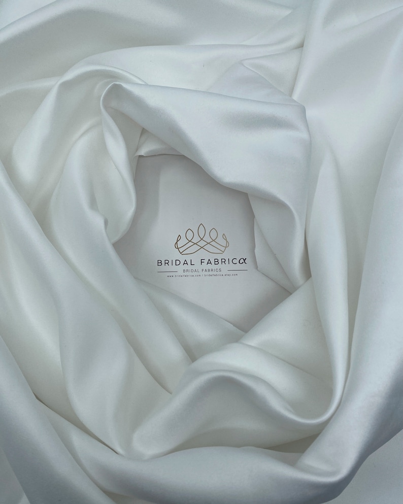 May include: White satin fabric, smooth and silky, draped in a soft curve. The fabric is perfect for wedding dresses, evening gowns, and other special occasion garments.  The fabric is available in a variety of widths and lengths.  Bridal Fabrica, Bridal Fabrics, www.bridalfabrica.com, bridalfabrica.etsy.com