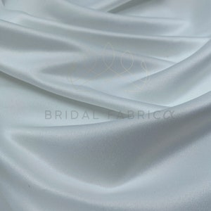 May include: White satin fabric with a smooth, silky texture. The fabric is draped and folded, showcasing its luxurious feel.