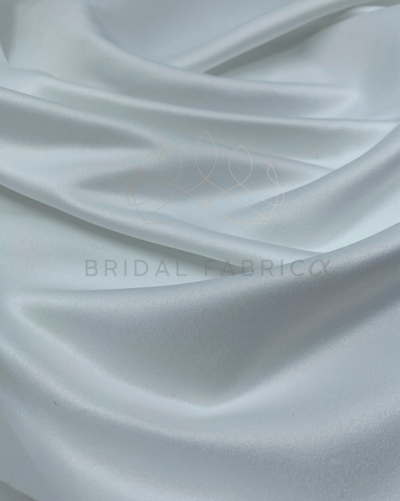 May include: White satin fabric with a smooth, silky texture. The fabric is draped and folded, showcasing its luxurious feel.