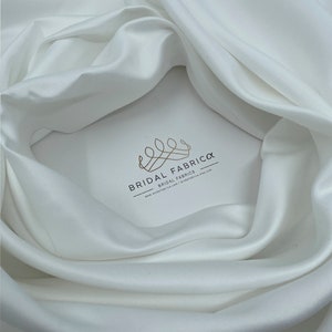 May include: White satin fabric with a soft, smooth texture. The fabric is draped in a way that shows its luxurious feel. The logo "Bridal Fabrica - Bridal Fabrics" is printed on the fabric.
