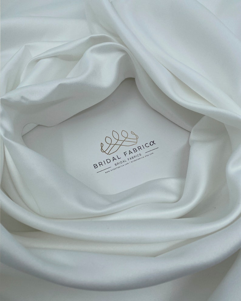 May include: White satin fabric with a soft, smooth texture. The fabric is draped in a way that shows its luxurious feel. The logo "Bridal Fabrica - Bridal Fabrics" is printed on the fabric.