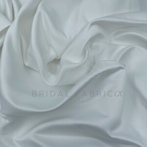 May include: White satin fabric with a smooth, silky texture. The fabric is draped and folded, showcasing its luxurious feel.