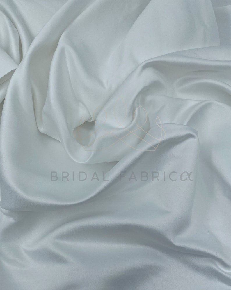 May include: White satin fabric with a smooth, silky texture. The fabric is draped and folded, showcasing its luxurious feel.