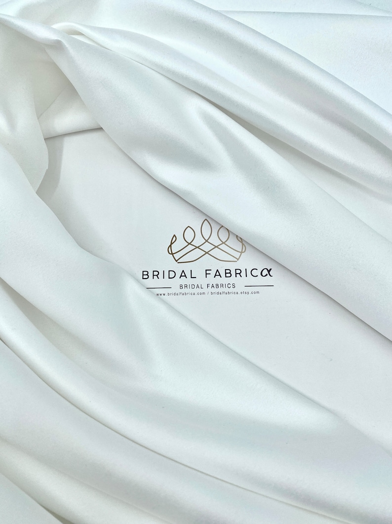 May include: White satin fabric, smooth and silky, with a subtle sheen. The fabric is draped in folds, showcasing its luxurious texture. The image includes the text "BRIDAL FABRICO" and "BRIDAL FABRICS" with a website address.