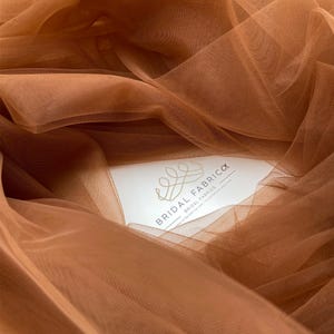 Può includere: Primo piano di un tessuto di tulle color ruggine con un cartoncino bianco su cui è scritto "Bridal Fabrccx Bridal Fabrics" e un logo a forma di corona.