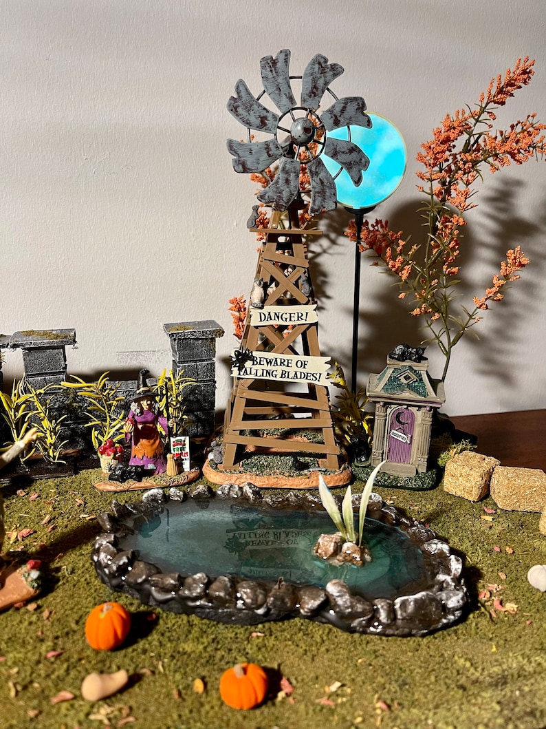 May include: A miniature Halloween village scene featuring a windmill, a pond, a small house, and a witch figure. The windmill has a sign that reads "Danger! Beware of Falling Blades!"