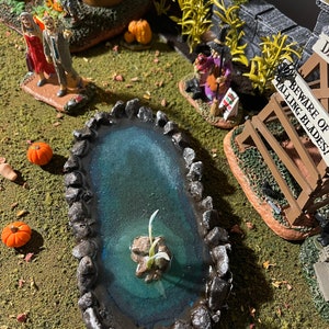 May include: A miniature Halloween scene featuring a small pond surrounded by rocks, a witch figurine, and a sign that reads "Beware of Falling Blades!"