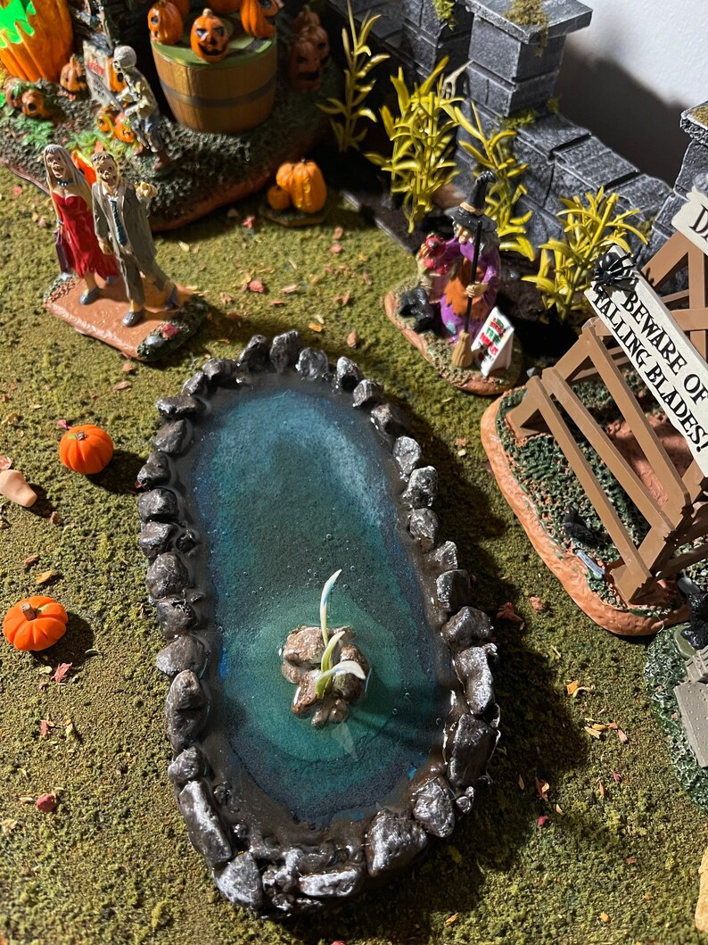 May include: A miniature Halloween scene featuring a small pond surrounded by rocks, a witch figurine, and a sign that reads "Beware of Falling Blades!"
