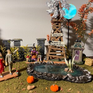 May include: A miniature Halloween village scene featuring a witch, a windmill, a pond, and a graveyard. The windmill has a sign that reads "Danger! Beware of Falling Blades!"