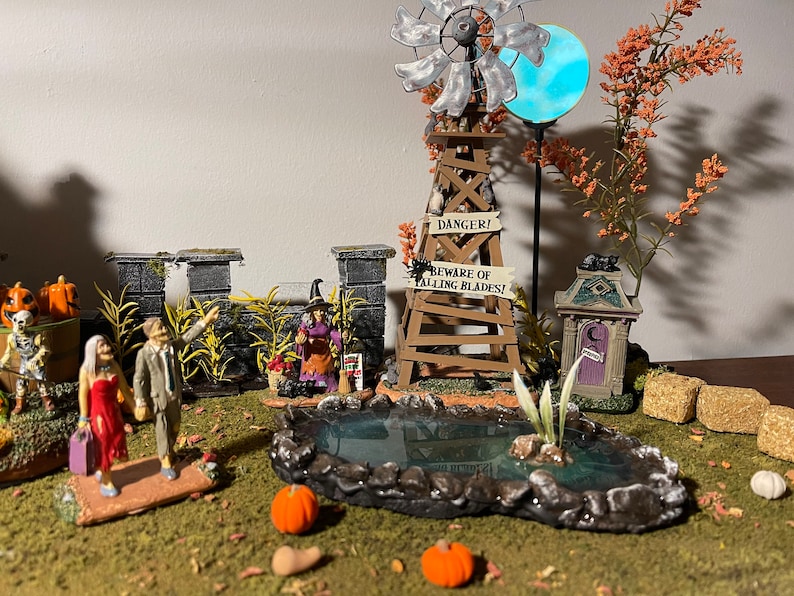 May include: A miniature Halloween village scene featuring a witch, a windmill, a pond, and a graveyard. The windmill has a sign that reads "Danger! Beware of Falling Blades!"