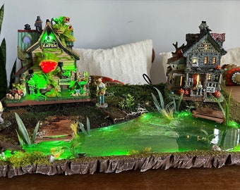 Könnte beinhalten: Eine Miniatur-Halloween-Dorfszene mit zwei Häusern, einem grünen Teich und einer Holzbrücke. Das erste Haus ist ein grünes Holzgebäude mit einem Schild, auf dem "The Shady Garden" steht. Das zweite Haus ist ein graues Holzgebäude mit einer Veranda und einem Schild, auf dem "Willkommen" steht.