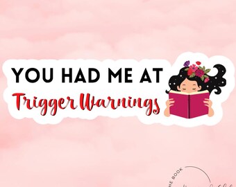 Może przedstawiać: Biała naklejka z czarnym tekstem, który brzmi "You had me at trigger warnings". Po prawej stronie naklejki znajduje się kreskówkowa ilustracja kobiety z długimi ciemnymi włosami i wieńcem kwiatowym, która czyta różową książkę. Tekst "The Book Boyfriend Club Est. 2022" znajduje się w kółku pod naklejką.