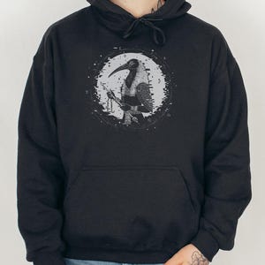 Op de afbeelding: Een zwarte hoodie met een grafische print van een pestmeesterfiguur binnen een cirkelvormig, verweerd wit kader. De hoodie heeft een kangoeroezak aan de voorzijde en bijpassende trekkoorden, gedragen met een bruine gebreide muts.