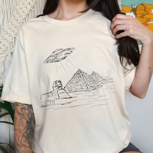 May include: A cream colored t-shirt with a black and white graphic of an alien spaceship hovering over the pyramids of Giza.