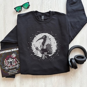 May include: A black crewneck sweatshirt featuring a white graphic of the Egyptian deity Thoth with a glitch effect, surrounded by a circular border. The sweatshirt is accompanied by black sunglasses and black over-ear headphones.