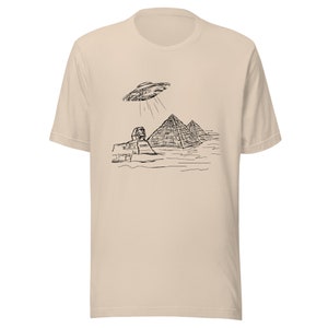 May include: Beige t-shirt featuring a black line drawing of the Sphinx, pyramids, and a UFO. The graphic depicts the UFO shining light beams down on the ancient Egyptian structures. The shirt has short sleeves and a crew neck.