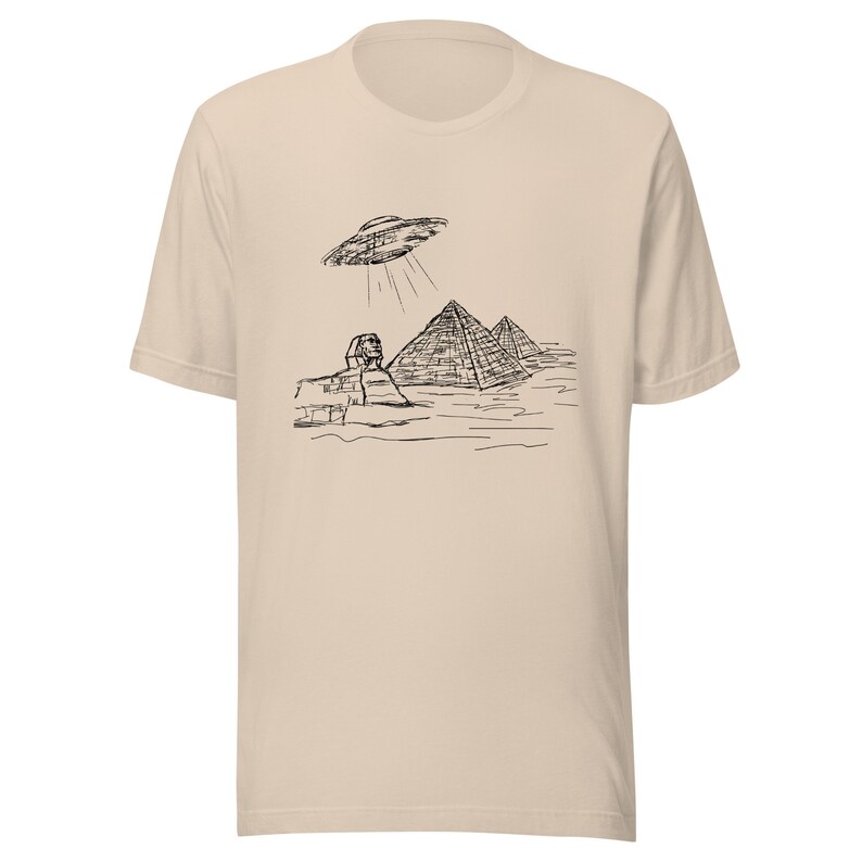 May include: Beige t-shirt featuring a black line drawing of the Sphinx, pyramids, and a UFO. The graphic depicts the UFO shining light beams down on the ancient Egyptian structures. The shirt has short sleeves and a crew neck.