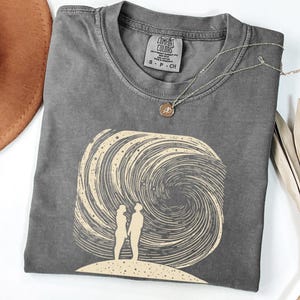 Puede incluir: Una camiseta gris con un estampado gráfico de dos figuras de pie en una colina con una galaxia giratoria detrás de ellas. El texto "COMFORT COLORS" está impreso en la etiqueta.