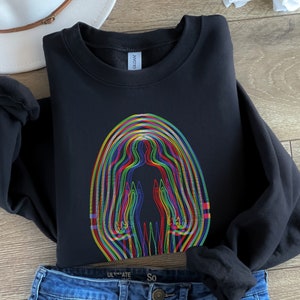 May include: Black crewneck sweatshirt featuring a colorful, layered human figure design. The graphic uses a spectrum of colors, including red, yellow, green, and blue. The sweatshirt is paired with blue denim jeans and a white hat.