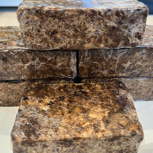 May include: A stack of six dark brown, rectangular bars of African black soap, wrapped in clear plastic. The soap has a textured surface and a natural, earthy appearance. The bars are stacked neatly, showcasing the product.