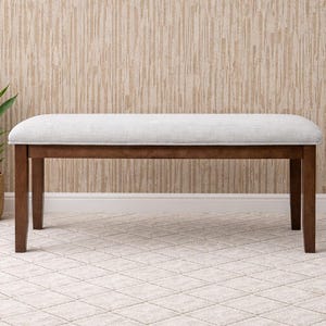 Puede incluir: Un banco rectangular con asiento tapizado gris claro y patas de madera marrón oscuro. El banco está colocado frente a una pared beige texturizada y se asienta sobre una alfombra estampada. Una planta en maceta está a la izquierda.