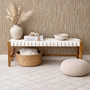 May include: A wooden bench with a white woven seat, holding decorative items. A woven basket, a ceramic vase with dried flowers, books, and a round knitted ottoman are also present. The background features a textured, vertical-striped wall.