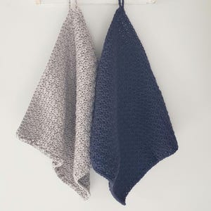 May include: Two hanging crochet towels, one in a light gray and the other in a deep navy blue. Each towel features a textured, knotted stitch pattern and a small loop at the top for hanging. The towels are displayed side by side against a plain wall.