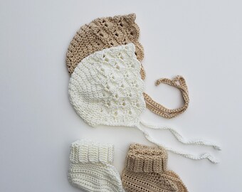 May include: Hand-crocheted baby bonnets and booties in cream and tan. The bonnets have scalloped edges and tie closures. The booties are ribbed at the ankle and have long ties. These handmade items are perfect for a baby shower gift.