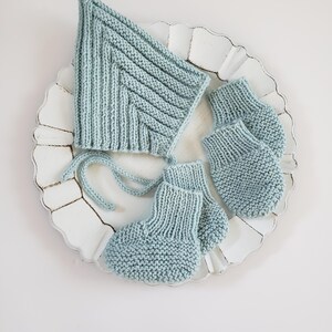 May include: A light blue knitted baby set, including a bonnet and two pairs of booties, displayed on a white, scalloped-edge plate. The bonnet has a pointed top and ties. The booties are small and cozy, perfect for a newborn.