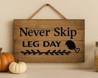以下が含まれることがあります： 「Never Skip LEG DAY」の文字が黒で書かれた木製の看板。ロープのハンガーと鶏の脚の黒いグラフィックが付いています。2つのカボチャと穀物の瓶が前景にあります。