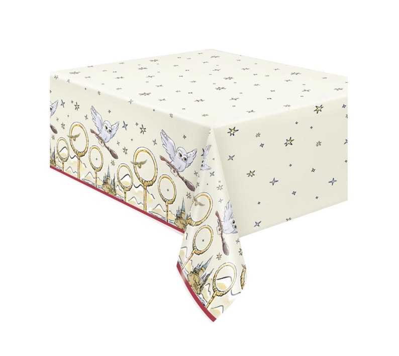 May include: Cream tablecloth with a repeating pattern of owls on brooms, golden hoops, and stars. The border has a maroon trim and a detailed illustration of a castle. A decorative table covering for themed events.