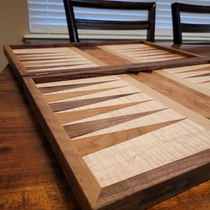 May include: A wooden backgammon board with a dark brown frame and a playing surface featuring alternating light and dark wood triangles. The board is open, revealing the playing field. The wood grain is visible, adding texture to the design.