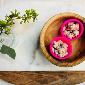 May include: Two pink silicone bowls filled with small, light brown star-shaped snacks, sitting inside a wooden bowl on a white marble surface.