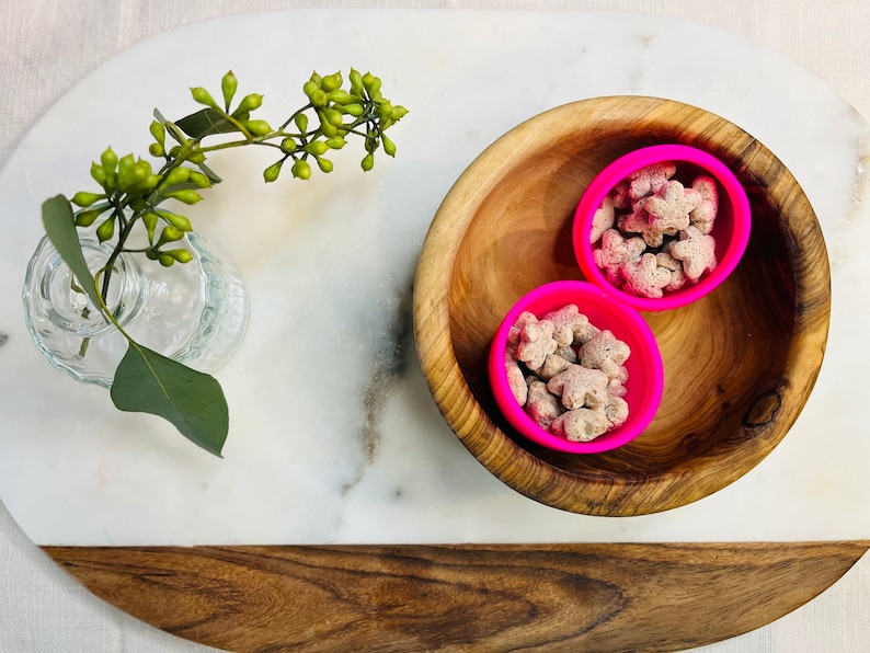 May include: Two pink silicone bowls filled with small, light brown star-shaped snacks, sitting inside a wooden bowl on a white marble surface.