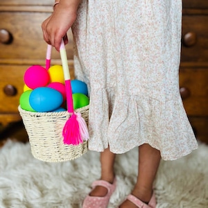 May include: A child holds a wicker basket filled with colorful plastic Easter eggs. The basket has a pink and white tassel hanging from it. The child is wearing a floral dress and pink shoes.