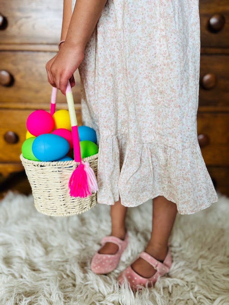 May include: A child holds a wicker basket filled with colorful plastic Easter eggs. The basket has a pink and white tassel hanging from it. The child is wearing a floral dress and pink shoes.