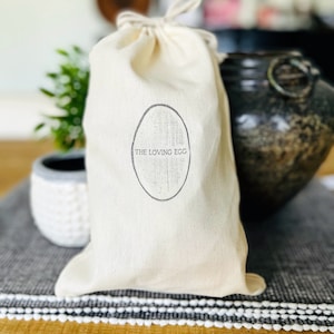 May include: A white canvas bag with a drawstring closure. The bag has a black oval logo with the text "THE LOVING EGG" printed inside.