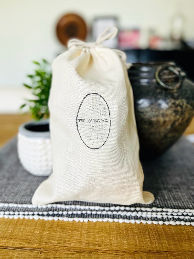 May include: A white canvas bag with a drawstring closure. The bag has a black oval logo with the text "THE LOVING EGG" printed inside.