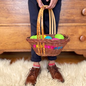 May include: A child holds a woven basket filled with colorful Easter eggs. The basket has a brown and tan woven design with a light brown handle. The child is wearing blue pants and brown shoes.
