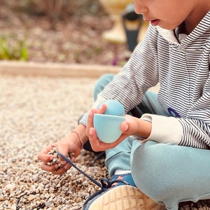 May include: A young person sits on a gravel surface, holding two light blue nesting cups. The person is wearing a striped hooded sweatshirt and blue pants.