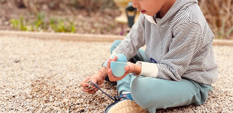 May include: A young person sits on a gravel surface, holding two light blue nesting cups. The person is wearing a striped hooded sweatshirt and blue pants.