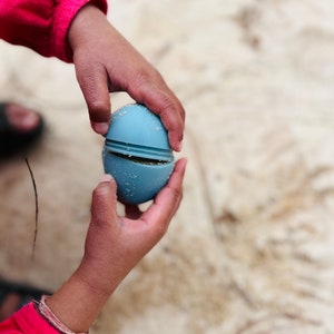 May include: A child's hand holds a blue silicone egg-shaped toy that is split in half. The toy is made of two halves that fit together.