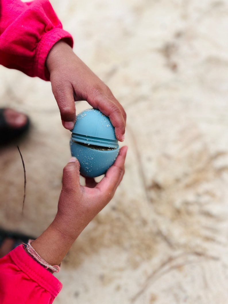 May include: A child's hand holds a blue silicone egg-shaped toy that is split in half. The toy is made of two halves that fit together.