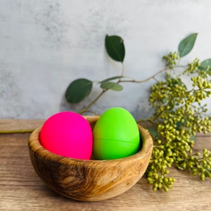 May include: Two colorful Easter eggs, one pink and one green, are nestled in a wooden bowl. The bowl has a natural wood grain pattern.