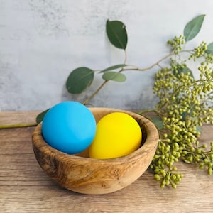 May include: A wooden bowl with a blue and yellow egg inside. The bowl is made of olive wood and has a natural finish.