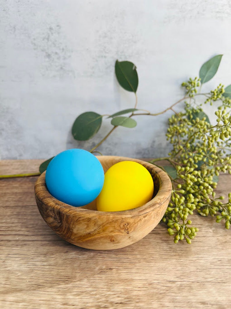May include: A wooden bowl with a blue and yellow egg inside. The bowl is made of olive wood and has a natural finish.