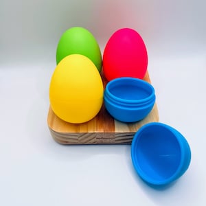 May include: Four colorful silicone Easter eggs on a wooden surface. Two of the eggs are closed, and two are open, revealing the inside of the eggs.