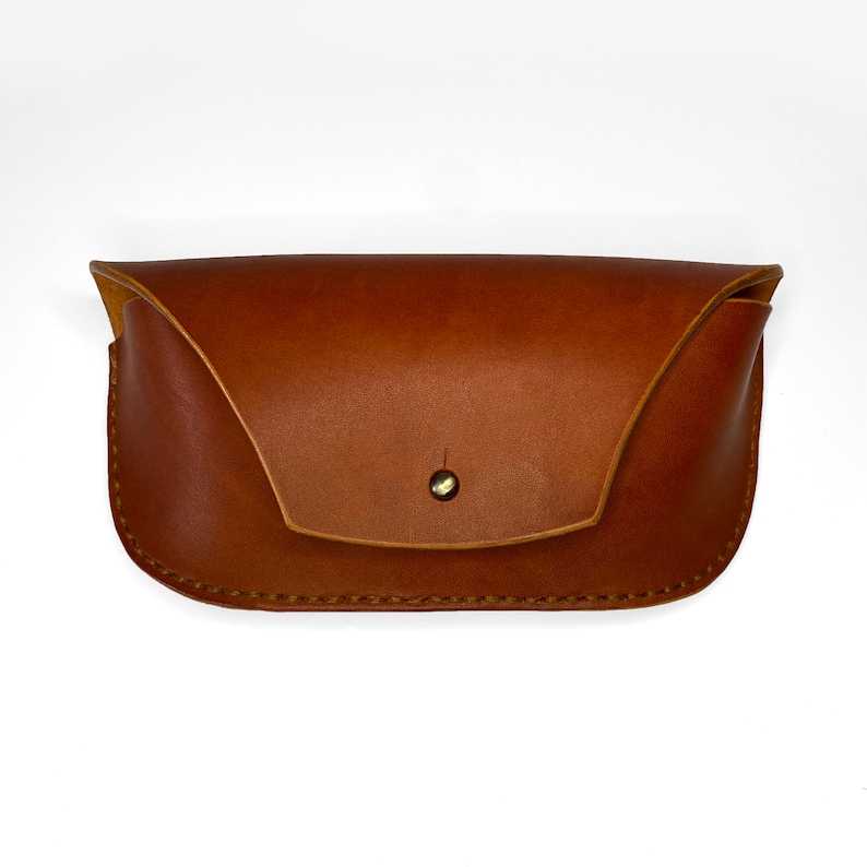 May include: A brown leather glasses case with a flap closure and a single button. The case is made of a soft, supple leather and has a subtle sheen.