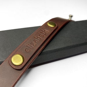 May include: A brown leather strap with gold metal accents and the name "elizabeth" embossed on it.